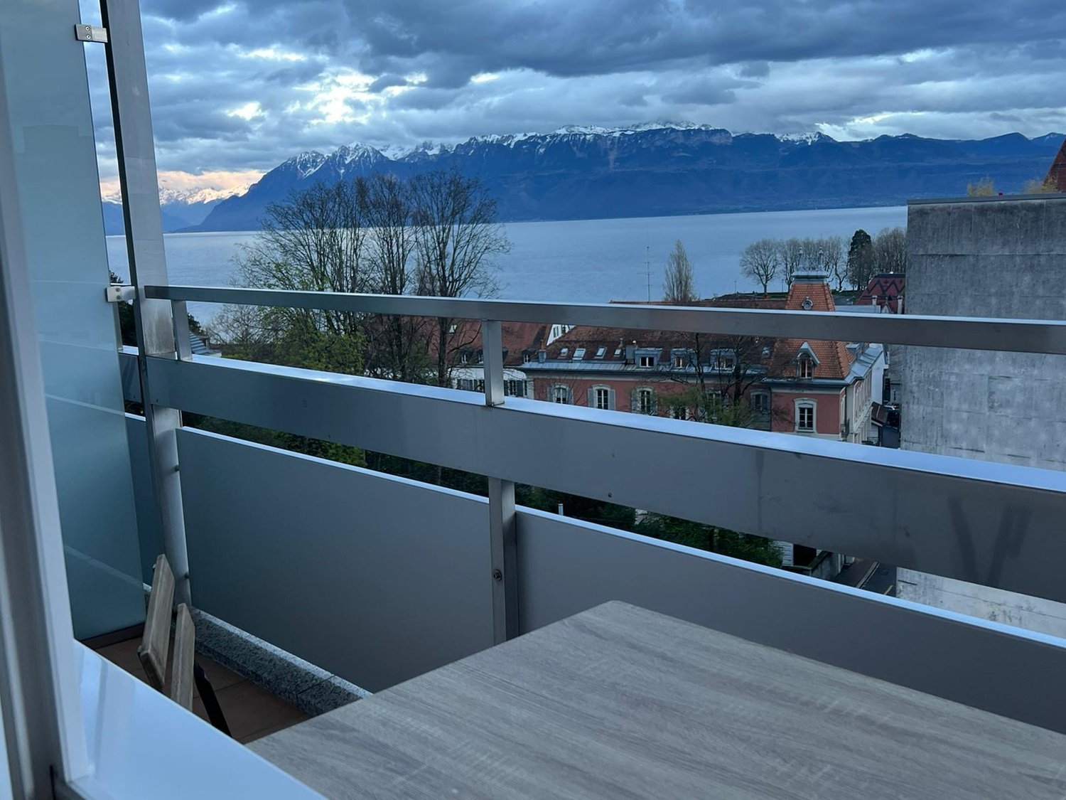 Balcony with table and chairs, metal railing, glass door, view of mountains and lake