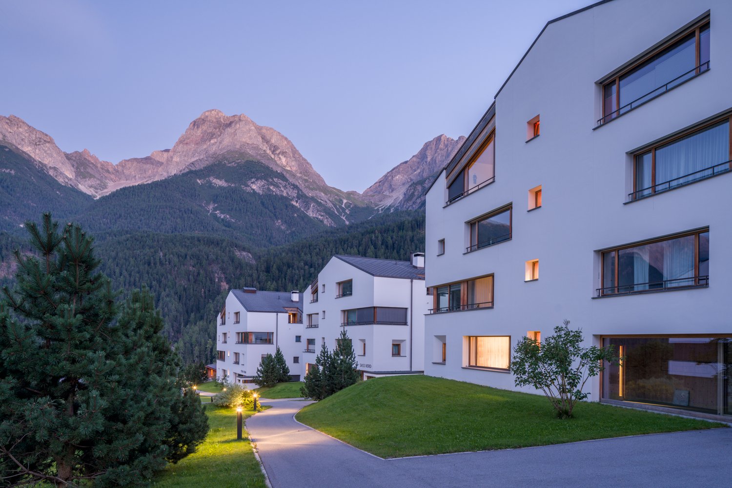 Modern apartment buildings with glass windows, concrete driveway, surrounding trees, green grassy area, mountain range at background