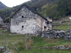 An old, dilapidated stone building in a rural, mountainous setting. The building appears to be in a state of disrepair, with crumbling walls and a partially collapsed roof. The surrounding landscape is lush and green, with mountains and hills in the backg