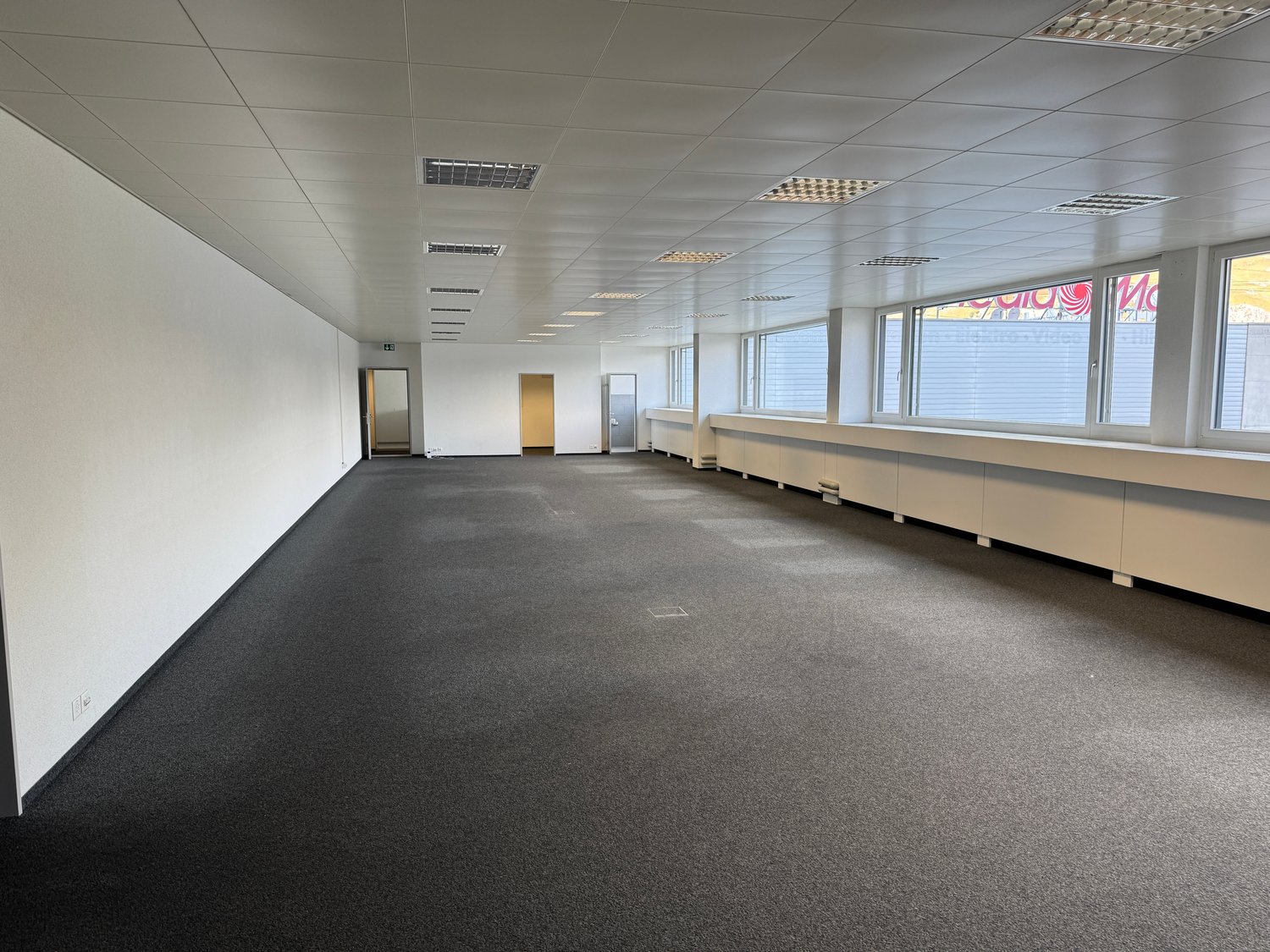 large empty room, gray floor, white walls, 2 doors, large windows, bench
