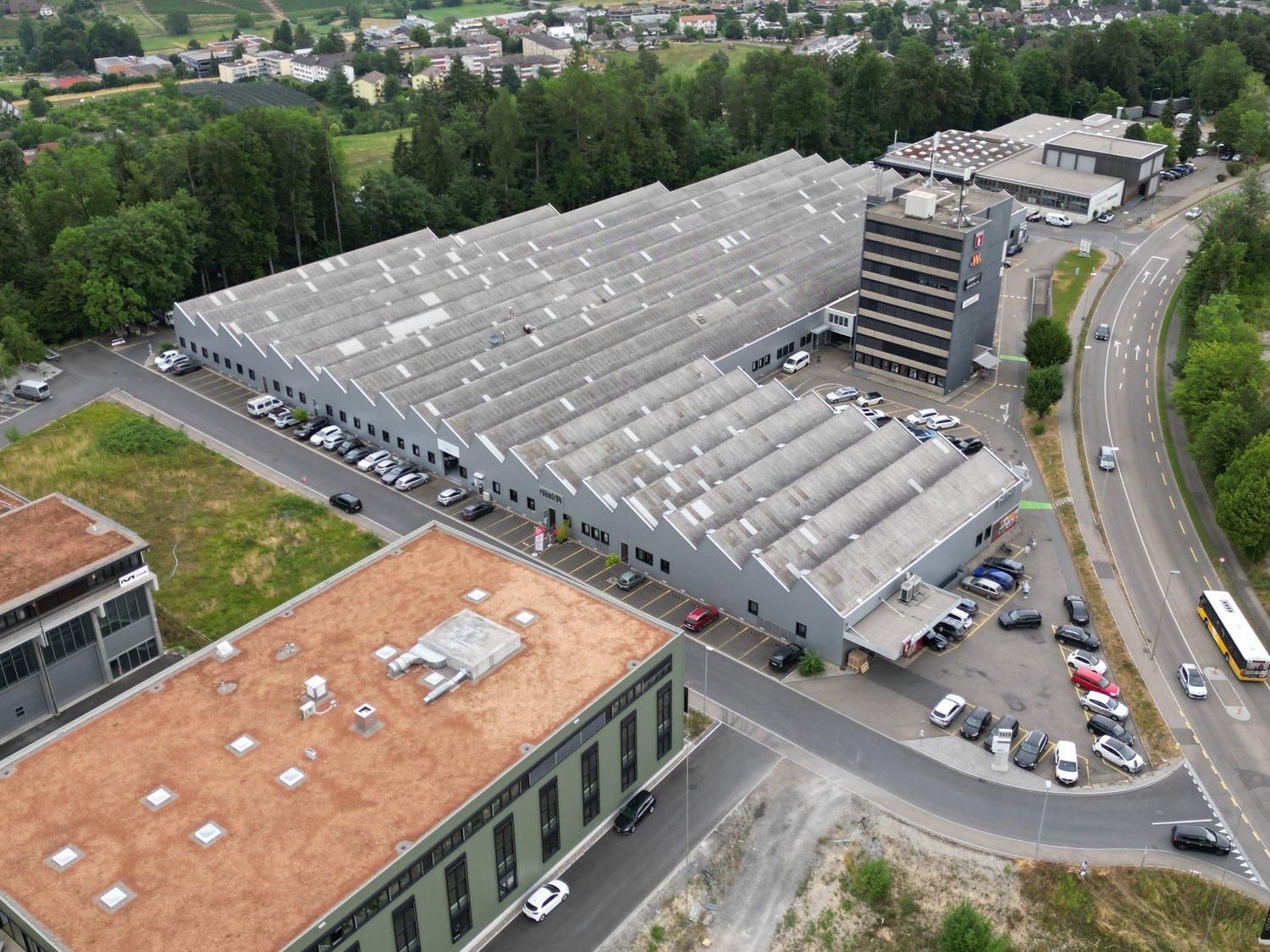 Large commercial building with a flat roof, multiple floors, and a parking lot in the foreground. The building appears to be an industrial or office complex surrounded by a residential area with trees and roads.