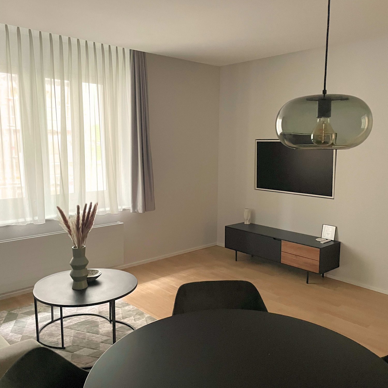A spacious living room with wooden floors and large windows, there is a modern hanging lamp above a round table, and a black sofa facing the TV mounted on the wall. A rug is placed in front of the sofa, and a side table holds a vase with tall plants. Behi