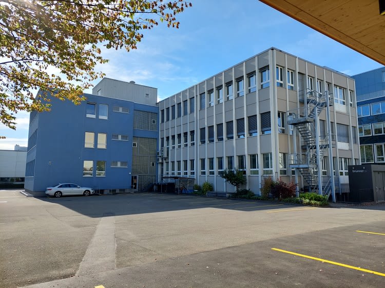 A multi-story office building with a modern, gray exterior. The building has numerous windows and appears to be located in an urban or industrial area with a parking lot in the foreground.