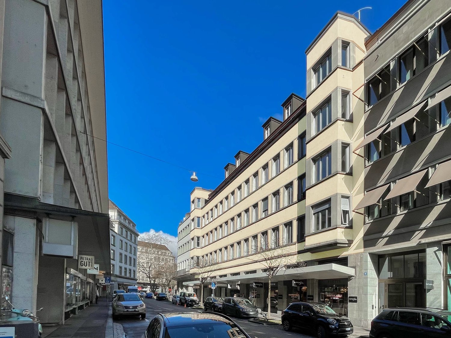 The image shows a street view of a city, with multi-story buildings lining the street. There are several parked cars visible on the street, indicating the presence of parking spaces. The buildings have a mix of architectural styles, with some having a mor