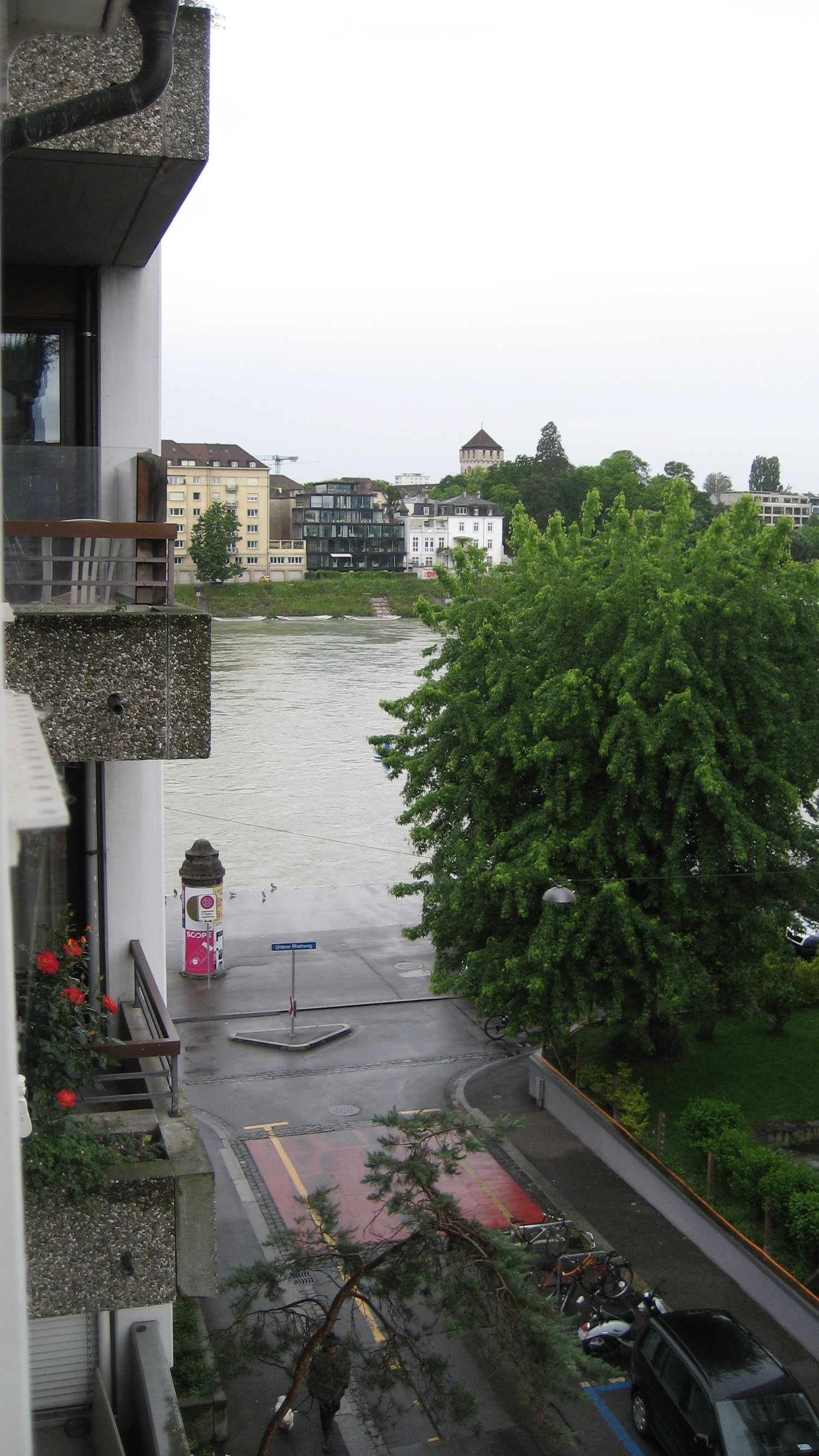 View of a city with a river, buildings, and trees. The image shows a rainy day with a gray sky and wet streets. There are various buildings, including some with towers, visible in the distance across the river.