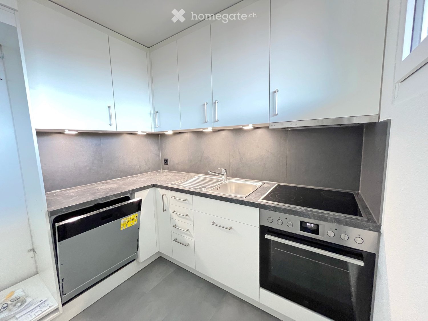 Modern kitchen with built-in dishwasher, oven, and washing machine. White cabinets, granite countertop, and sleek sink.