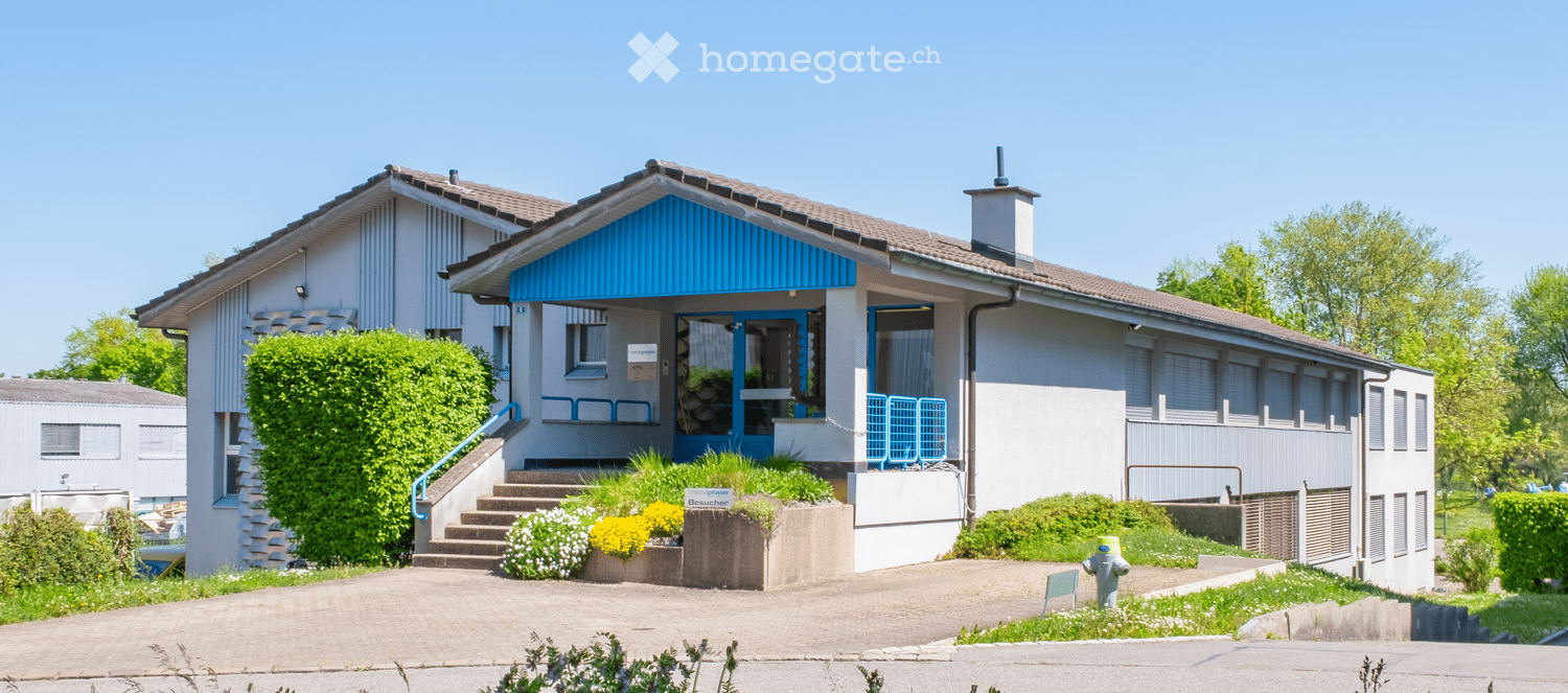 2-story house with white walls, blue roof, and a covered porch entrance. The house has a balcony or terrace on the second floor. There are green bushes and plants surrounding the house, and a paved driveway or walkway leading to the entrance.