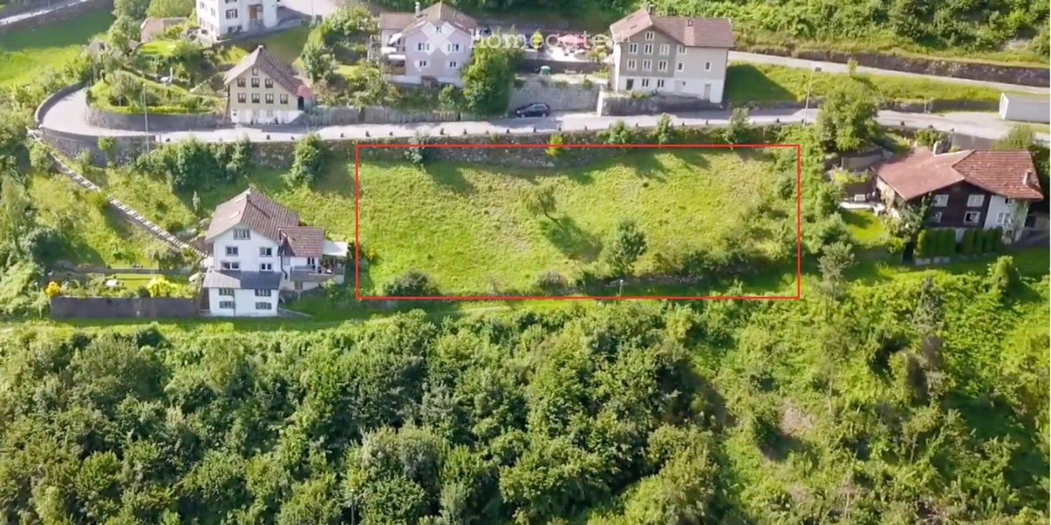 The image shows an aerial view of a residential area with several houses and buildings surrounded by lush greenery. There appears to be a paved road or path running through the area. Within the highlighted area, there is an open space that could potential
