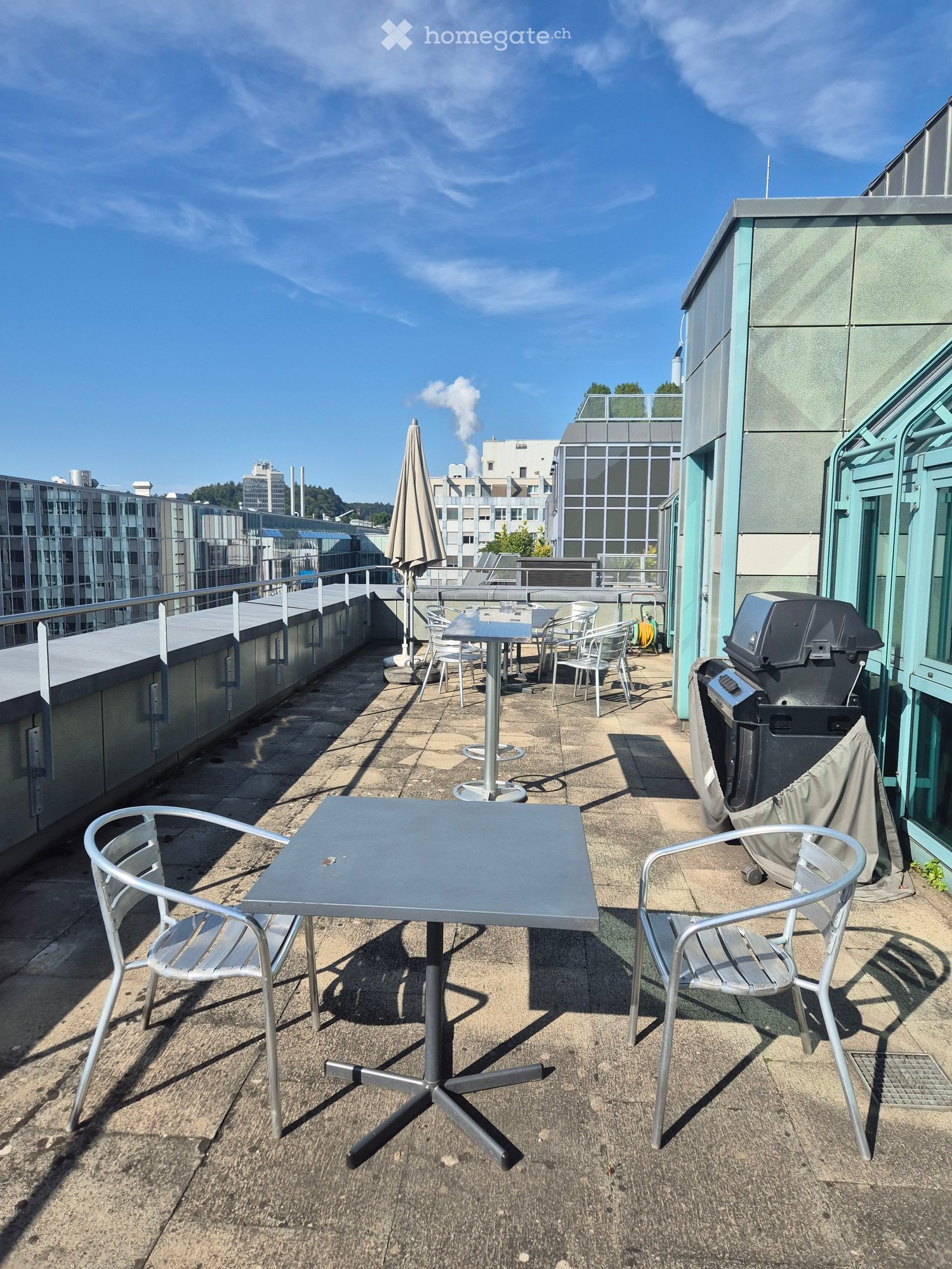 The image shows an outdoor terrace or balcony area with a table and chairs, overlooking a cityscape with buildings and a blue sky with wispy clouds. There is also a closed umbrella and some outdoor equipment visible.