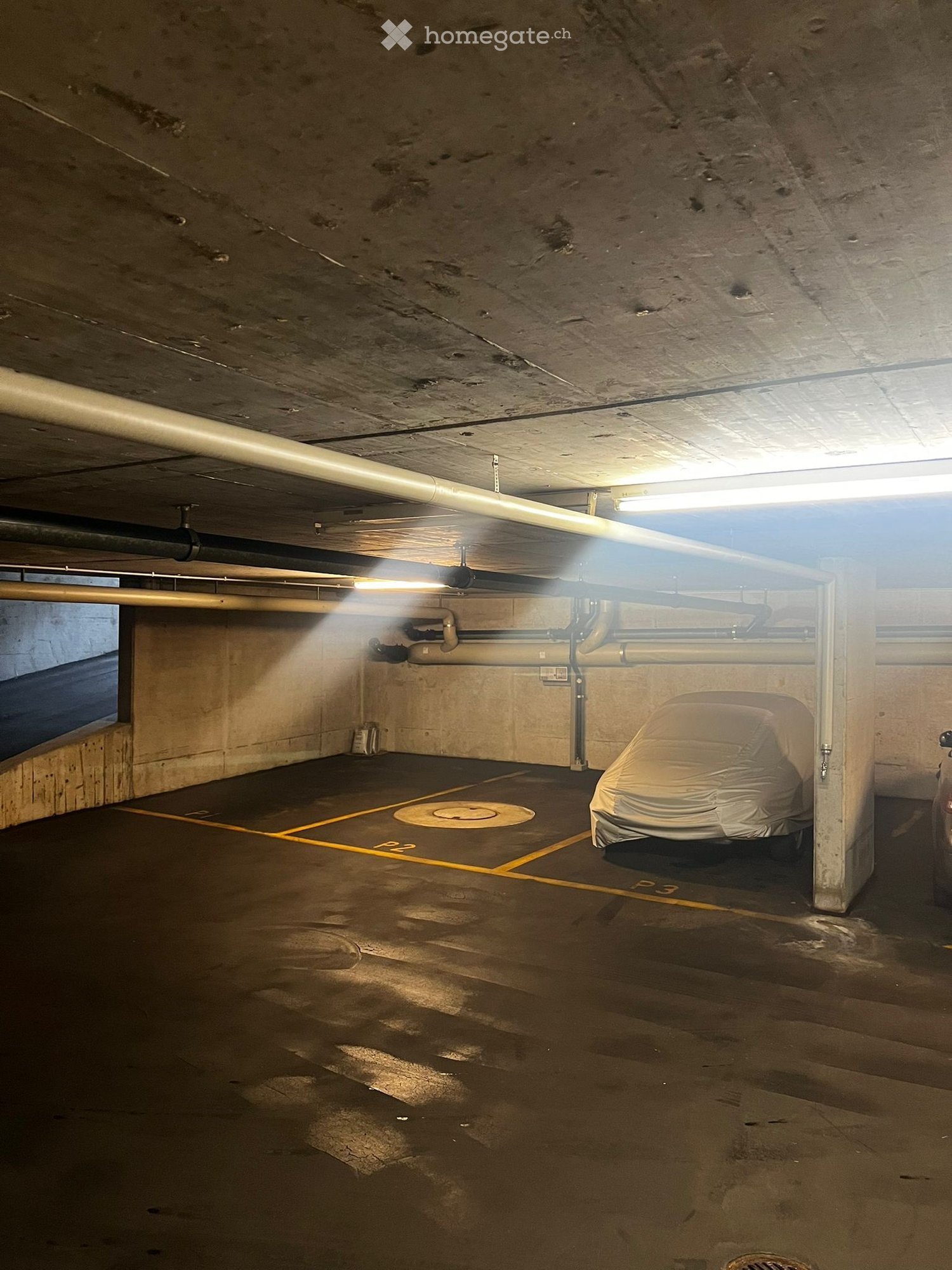 Dimly lit underground garage with concrete floor, pipes and wires running along the ceiling, and a covered vehicle parked in one of the spaces.