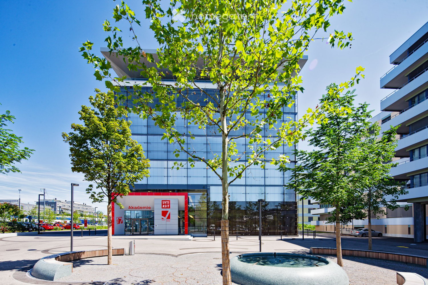 Modern glass and steel building with a large tree in the foreground, surrounded by a plaza with benches and a water feature. The building has a red sign that says 'SVTI ASIT Akademie'.