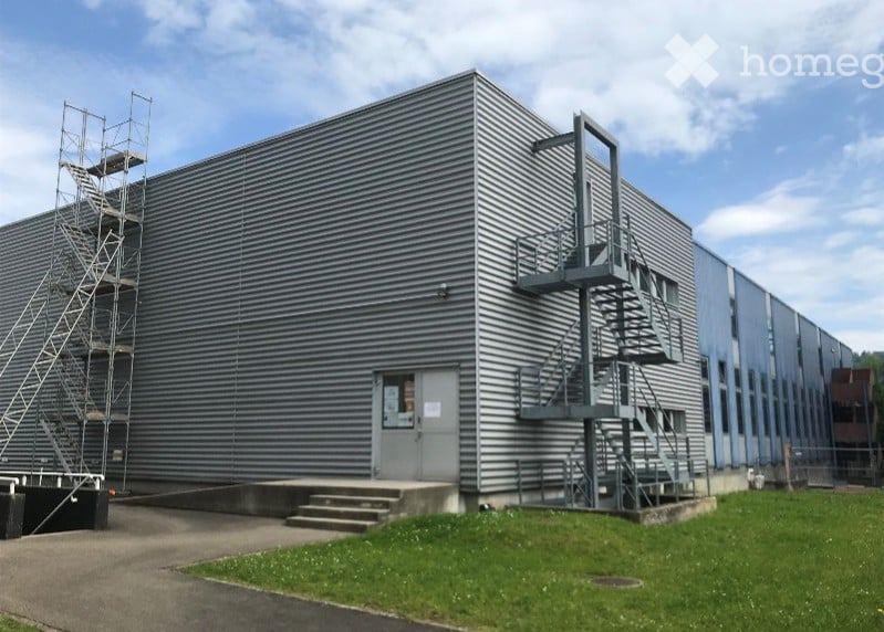 Large industrial building with a gray metal exterior, multiple floors, and an exterior staircase. There appears to be a parking area in front of the building.