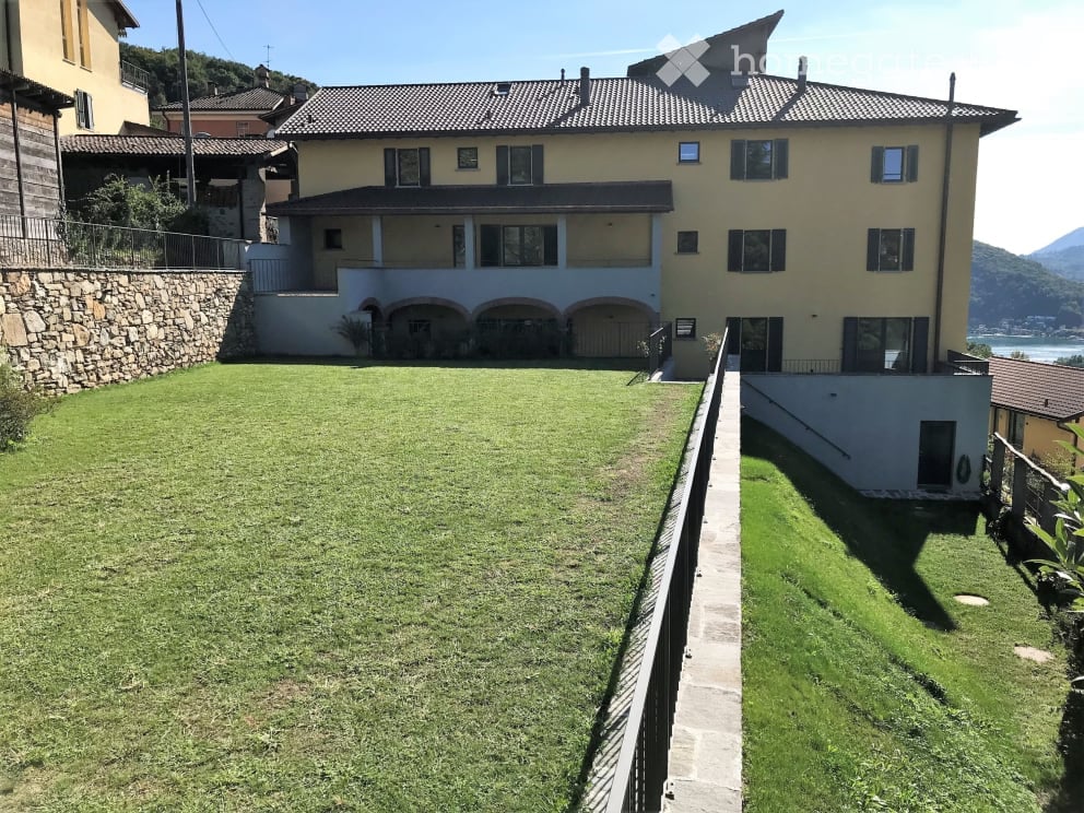 3-story apartment building with a stone facade, arched windows, and a grassy yard with a paved walkway leading to the building. The building appears to have balconies or terraces on some of the floors.