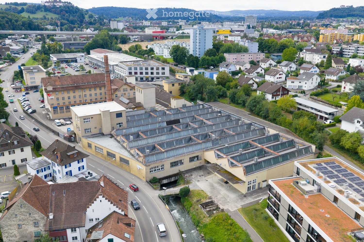 large building complex with multiple structures, some with solar panels, near a river and roadway, many surrounding houses and smaller buildings
