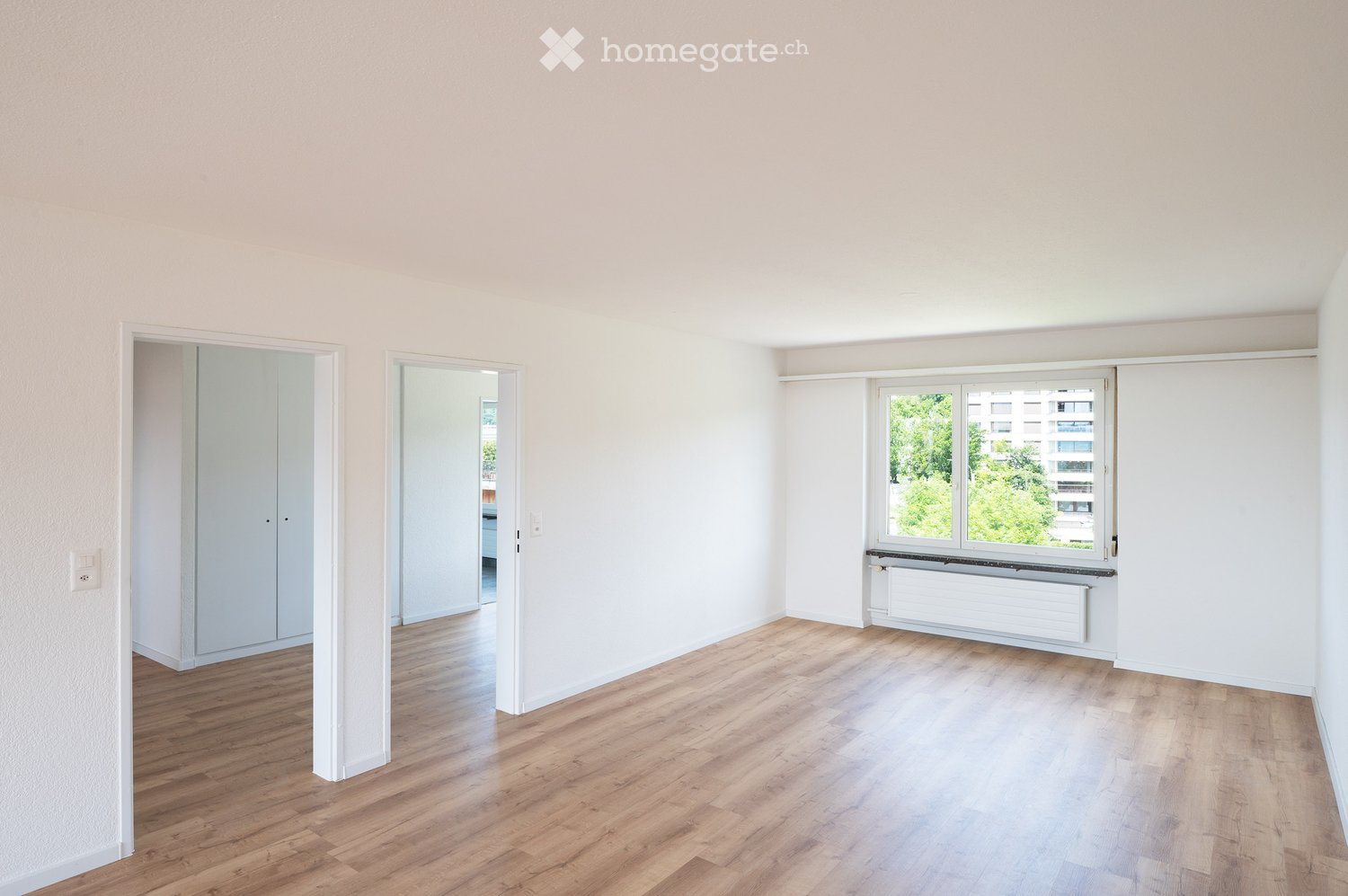 Spacious living room with large windows, hardwood flooring, and built-in closets. The room has a bright and airy feel with a view of the greenery outside.