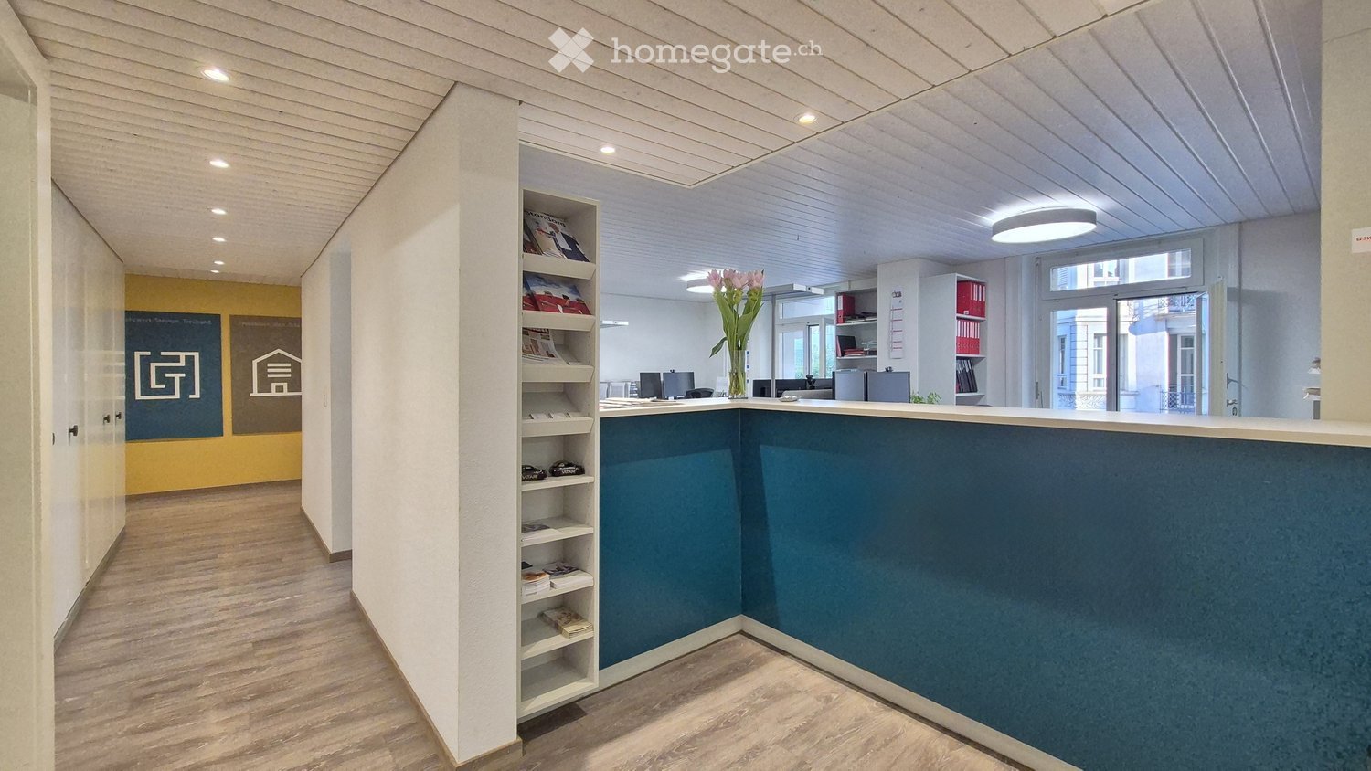 Modern office space with white walls, wooden floors, and built-in shelving. The reception area features a blue counter and storage cabinets. The space appears to be well-lit with recessed lighting and large windows.