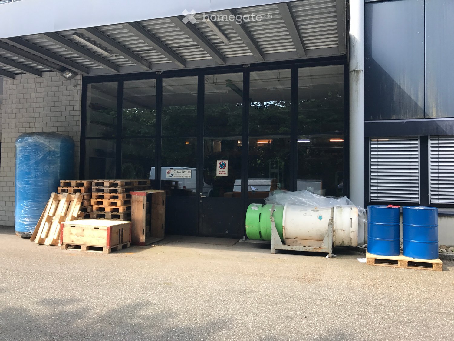 Metal building exterior with glass doors and windows, large entryway, blue barrels, green metal cylinders, wooden pallets, and blue plastic coverings. Bright daylight illuminating the area.