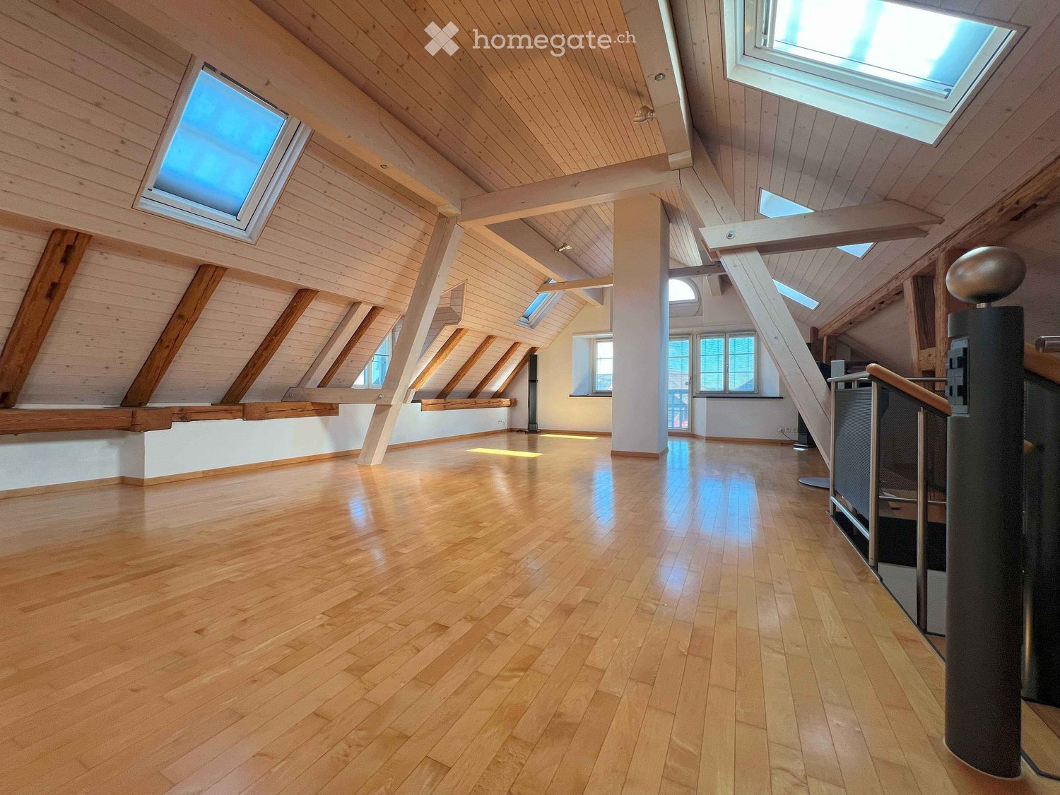 A spacious attic room with hardwood floors, wooden beams, multiple skylights, and a high ceiling. The room is well-lit with natural light coming through the windows. It has a modern and open design, with a minimalistic decor.
