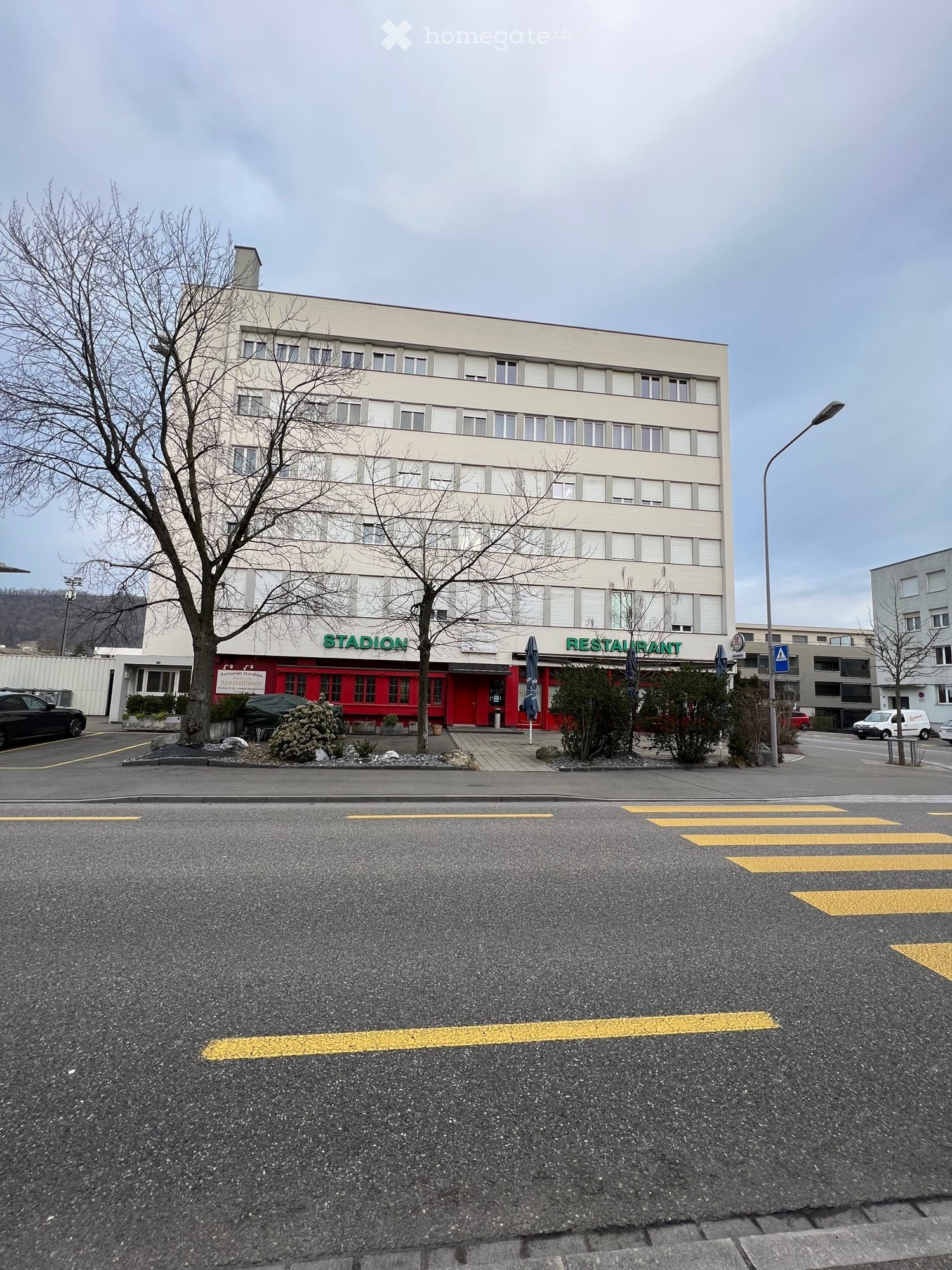 3-story building, white walls, red facade with text 'Stadion', restaurant with blue umbrellas, parking lot in front