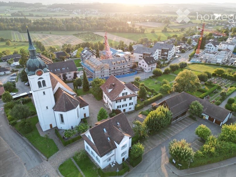 Aerial view of a town featuring a church, houses, a construction site, a bus, greenery, and buildings. A tower with a clock is visible on the church. A bus is visible on the street. Several parked cars are on the right side.