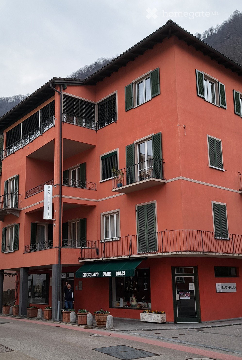 Red building with 4 floors, balcony, several windows, 4 planters with flowers, bakery shop.