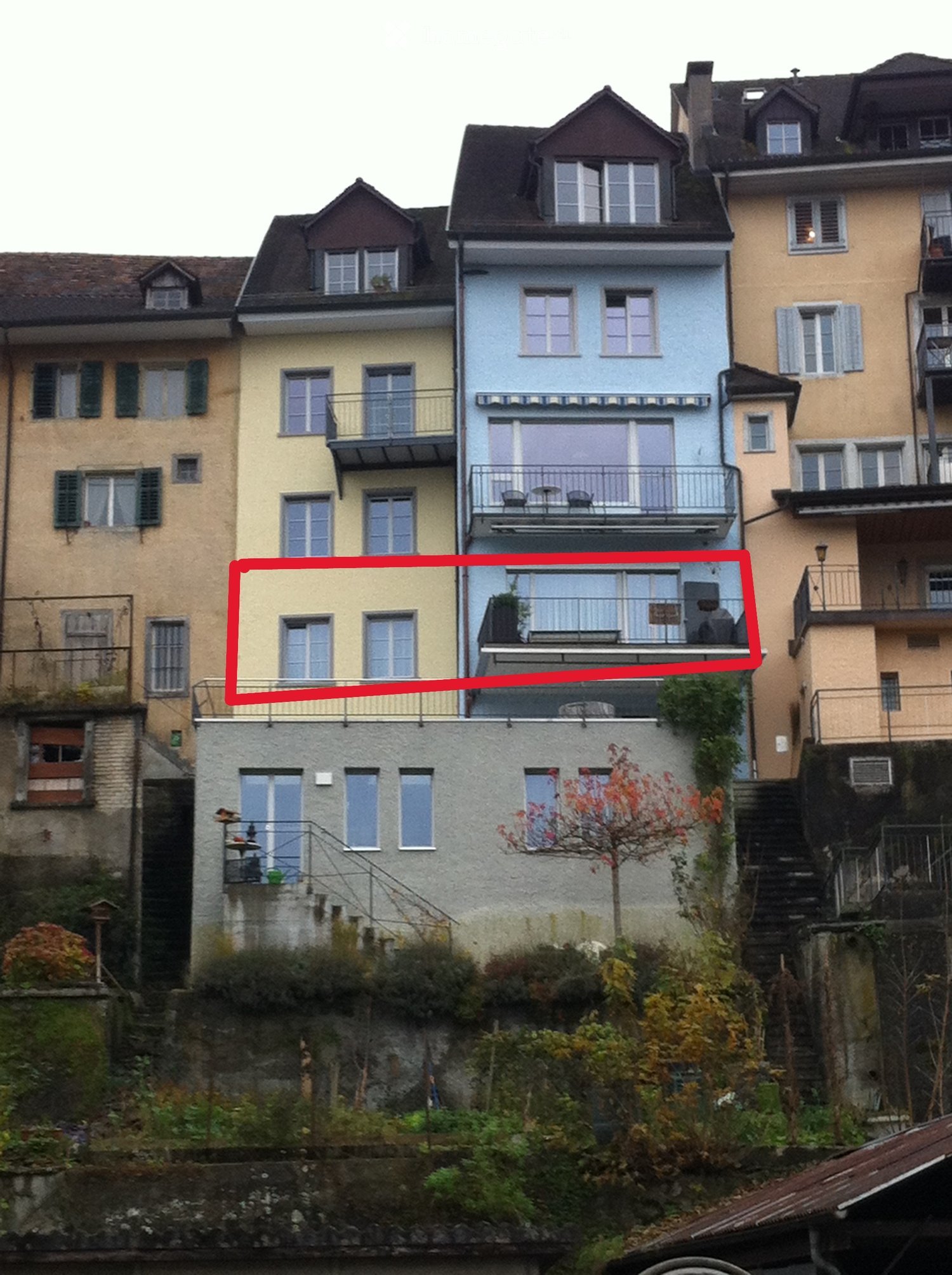 Residential building with multiple floors, yellow and blue exterior walls, multiple windows, balcony in the marked section