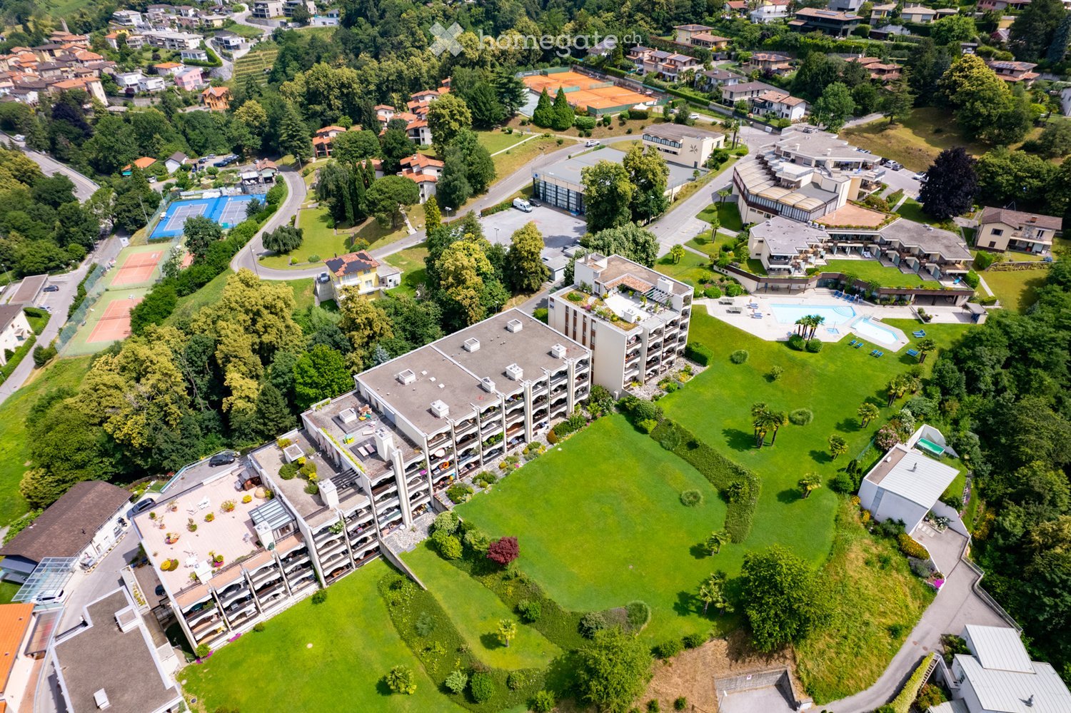 Complex with multiple buildings, swimming pool, parking lots, tennis courts, surrounded by green area