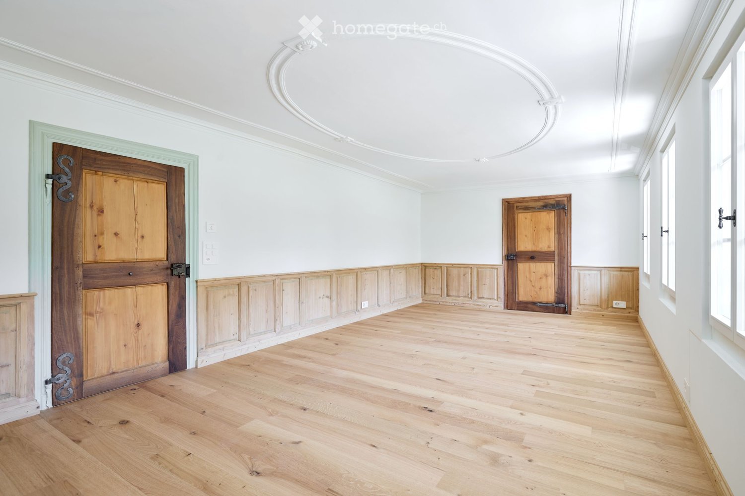large empty room, white walls, wooden doors, wooden flooring, large windows, molding on wall