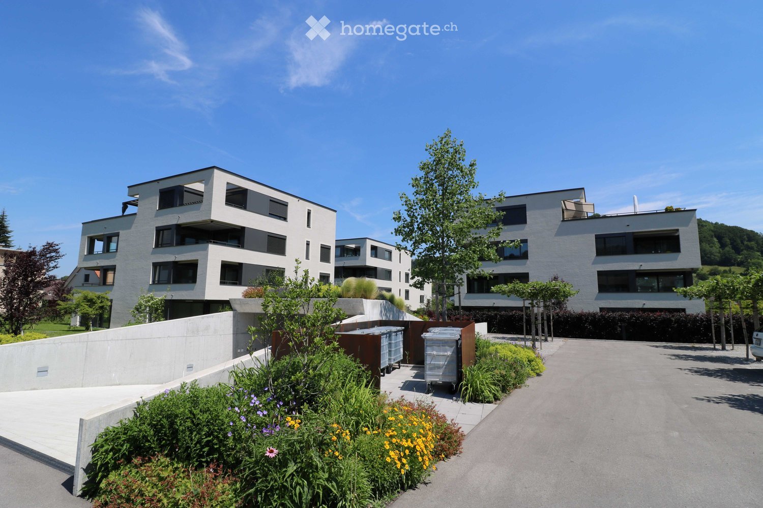 Three modern buildings with white exterior, balconies, and large windows. The area is surrounded by well-kept gardens, with shrubs and flowers.