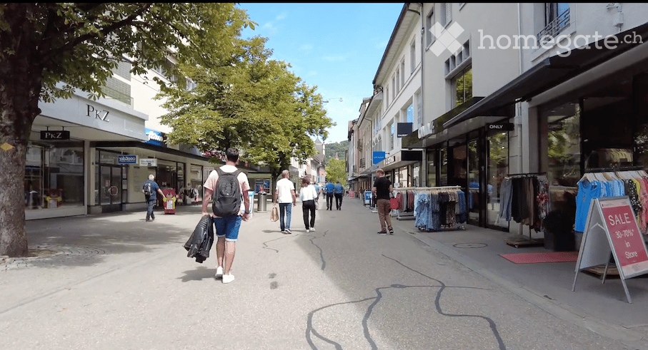 People walking on a shopping street, stores on both sides, trees and parked cars in the background.
