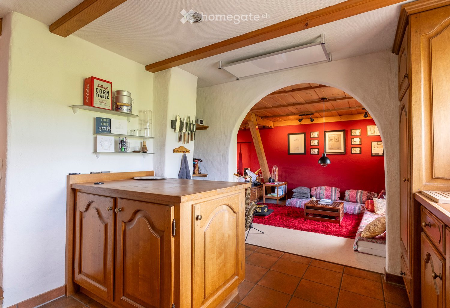 Wooden counter, cabinets, wooden shelves, kitchen tools, red wall, arched entrance, couch, table, pillows, red carpet, framed pictures