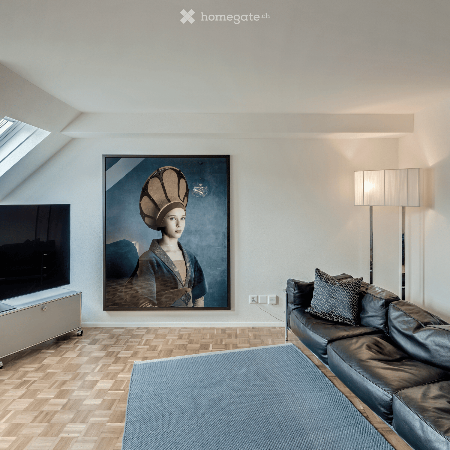 Living room with white walls, wooden floor, black couch, carpet, picture frame, floor lamp, wall socket, TV