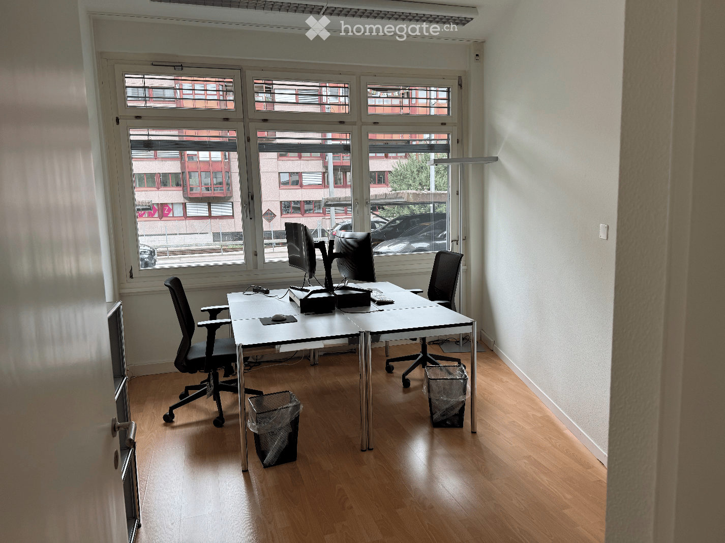empty office room with white walls, wooden floor, desk, 2 office chairs, 2 computer monitors, large windows, trash cans