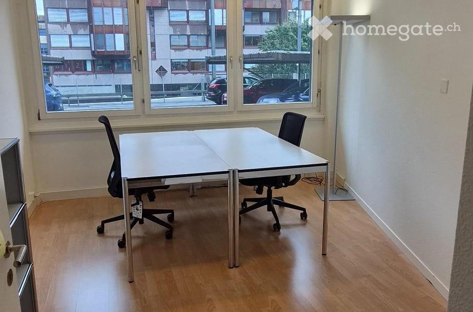 A small, unfurnished room with a white table and two black chairs, large windows showing a cityscape, and a modern floor lamp in the corner.
