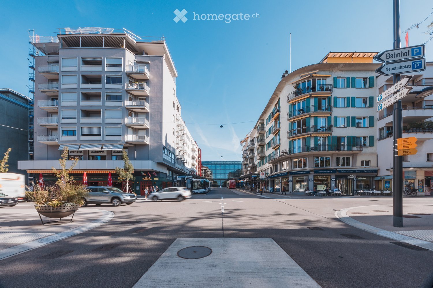 Urban street scene with multi-story buildings, balconies, storefronts, and traffic signs. Clear blue sky and modern architecture.