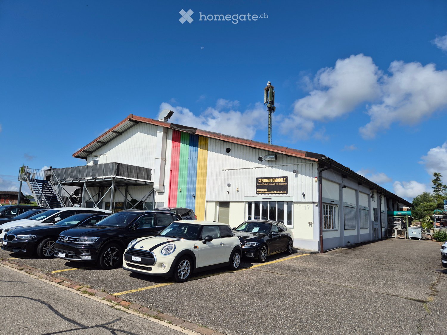 Warehouse with cars parked in front, white exterior, rainbow stripes, staircase, tower on the roof