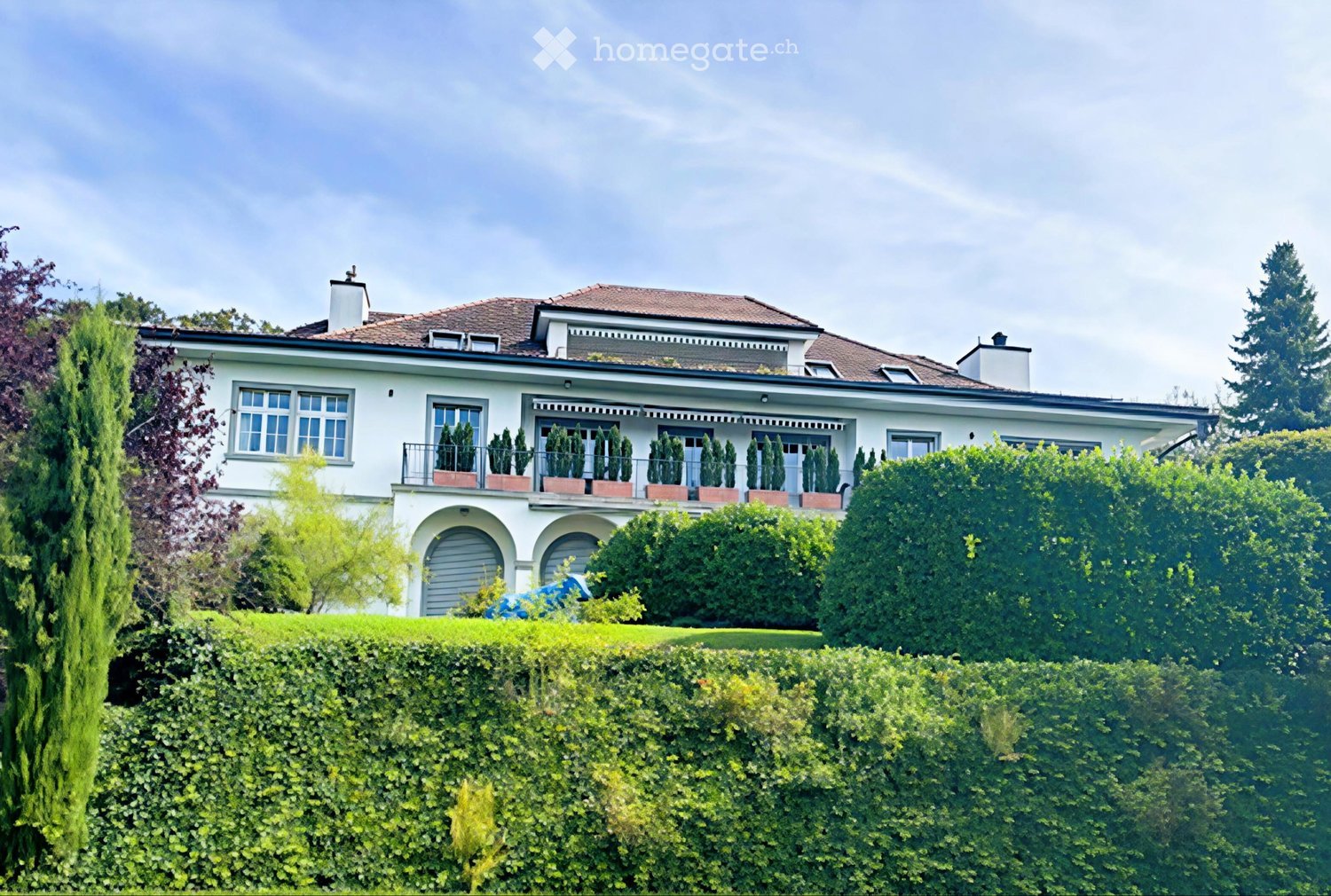 2 story white house with red roof, garage, balcony with plants, well-kept garden