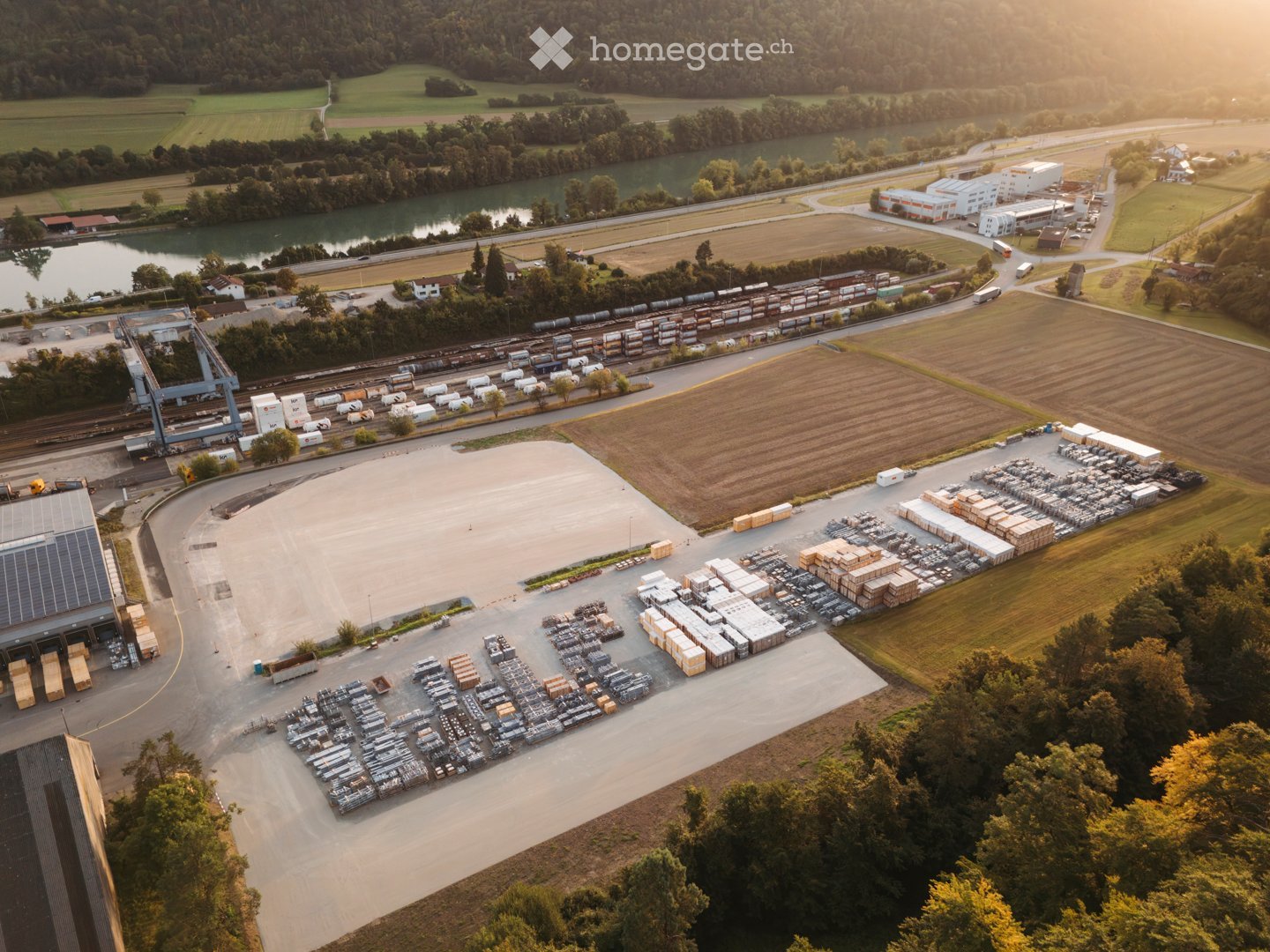 Aerial view of an industrial complex with storage facilities, a large open area, and various vehicles, including trucks and containers, near a river and railway line.