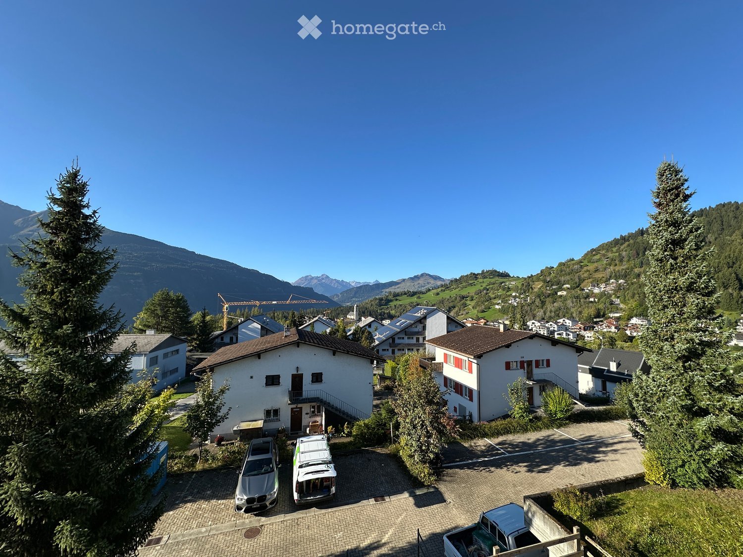 Multiple residential buildings with brown roofs, white walls, surrounded by greenery, clear blue sky, mountains in the background