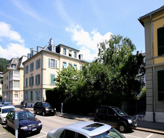 A large, beige building with many windows and a gray roof, with cars parked in front, near trees and bushes.