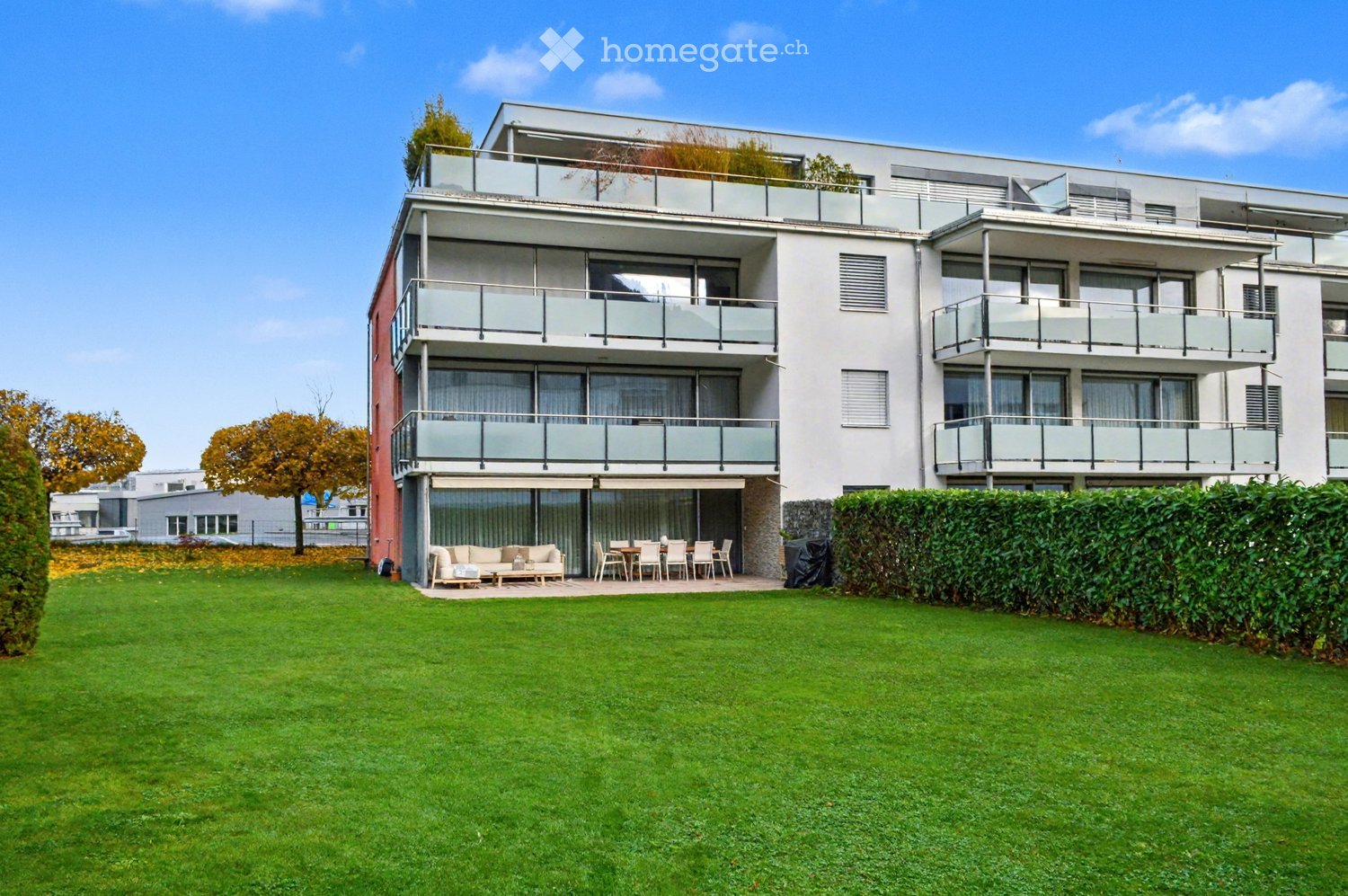 Modern building with 4 levels, each with balconies, front yard with green lawn and hedges, large windows, patio with outdoor table and chairs, garden furniture
