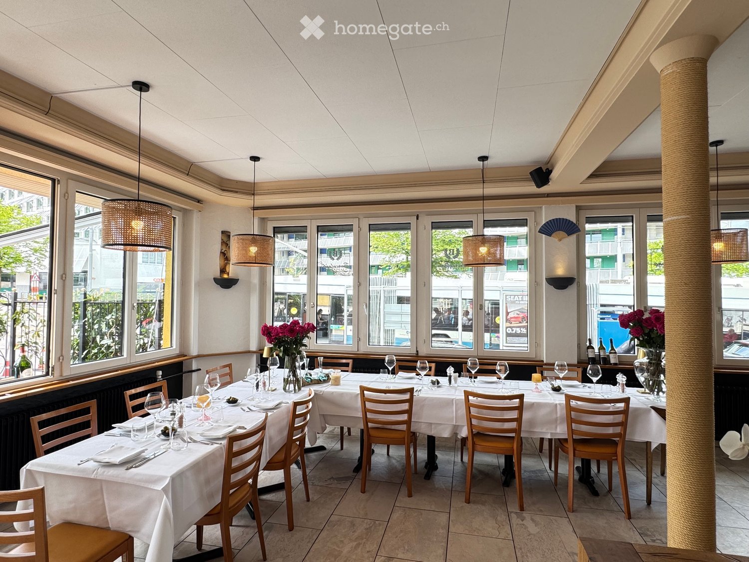 Dining area with two long tables, wooden chairs, wine glasses, plates, vases with red flowers, pendant lamps, and windows with large panes