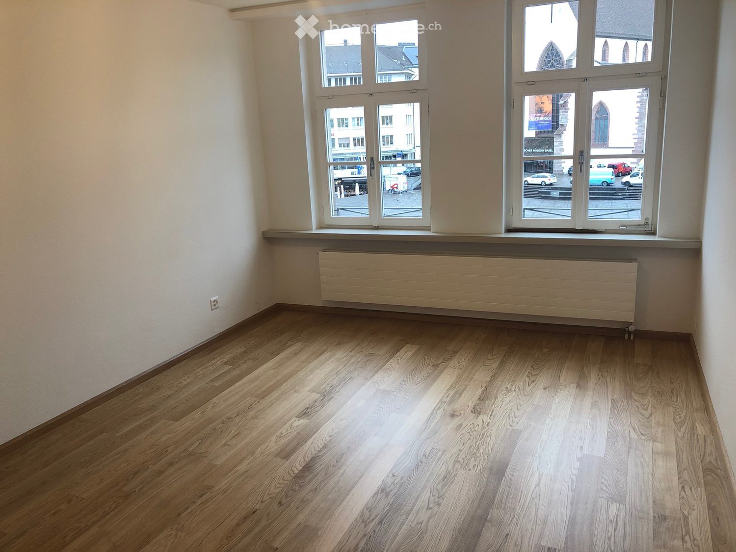 This image shows a spacious room with large windows overlooking a city street. The room has a hardwood floor and white walls. There is a built-in cabinet or shelf unit along one wall.