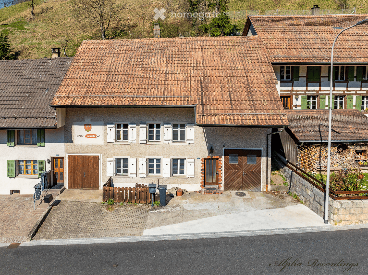 Aerial view of a house with a brown roof, white shutters, wooden doors, brick paving, garbage bins, and a lamp post.