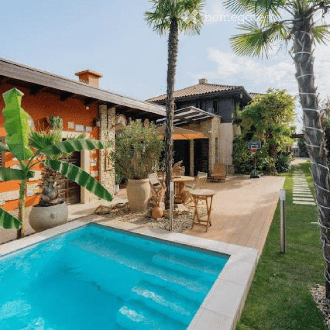 Swimming pool with clear blue water, grassy lawn, brick building, small house, large palm trees, potted plants, patio umbrella, garden table and chairs, basketball hoop