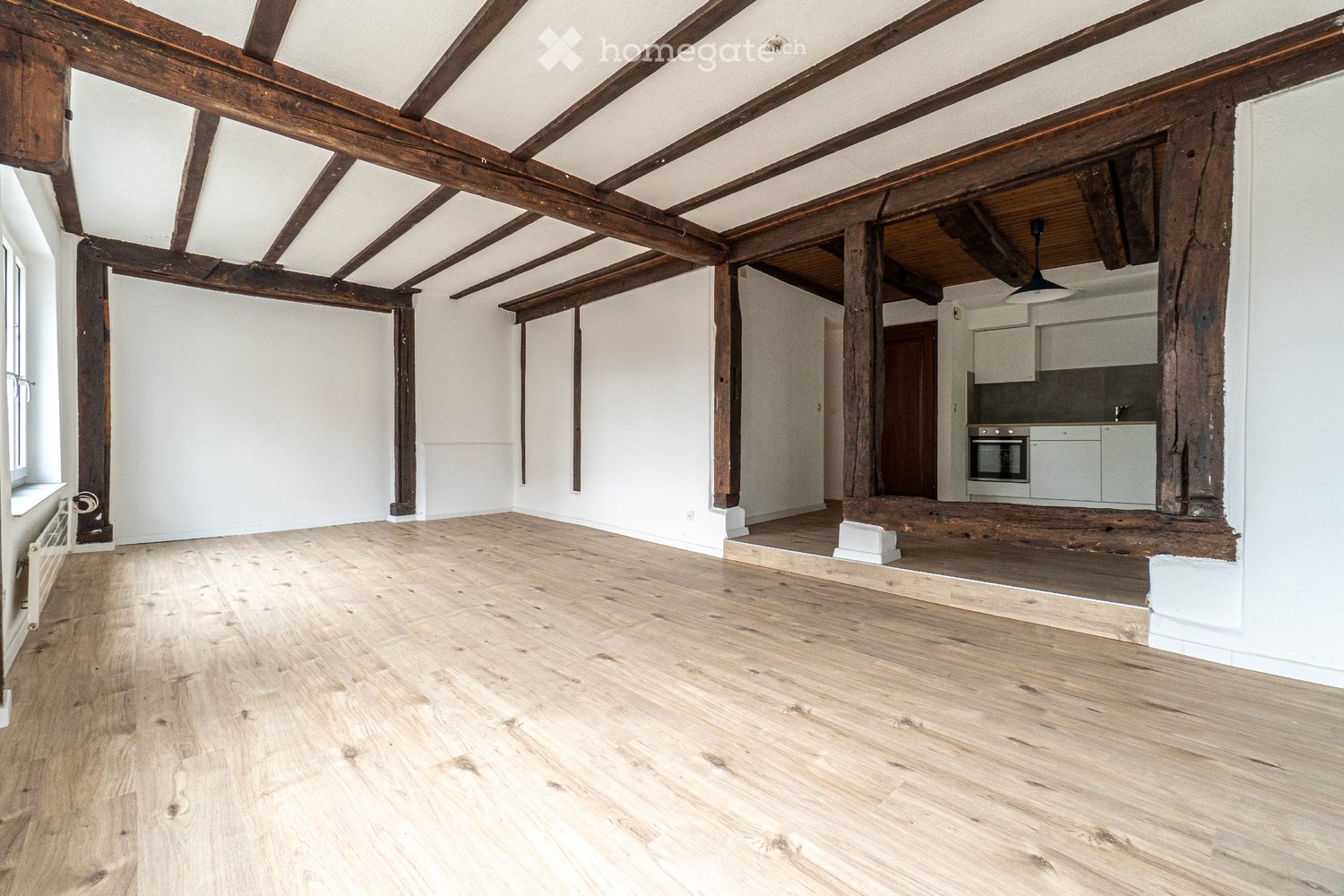 wood floor, exposed wooden beams, white walls, open view to kitchen, windows