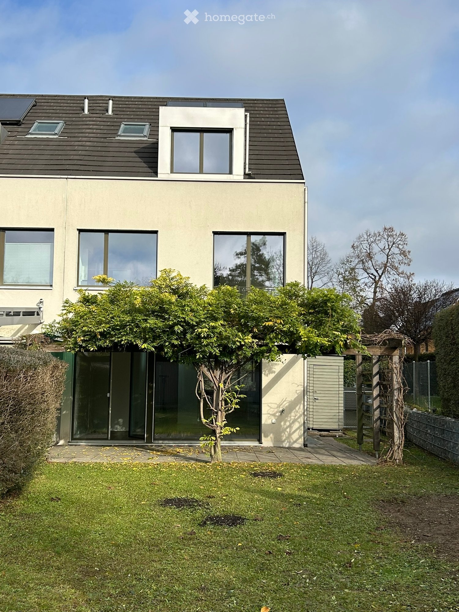 2 story house with large windows, grey exterior, solar panels on the roof, glass doors and windows, grass garden, shrub, pergola.