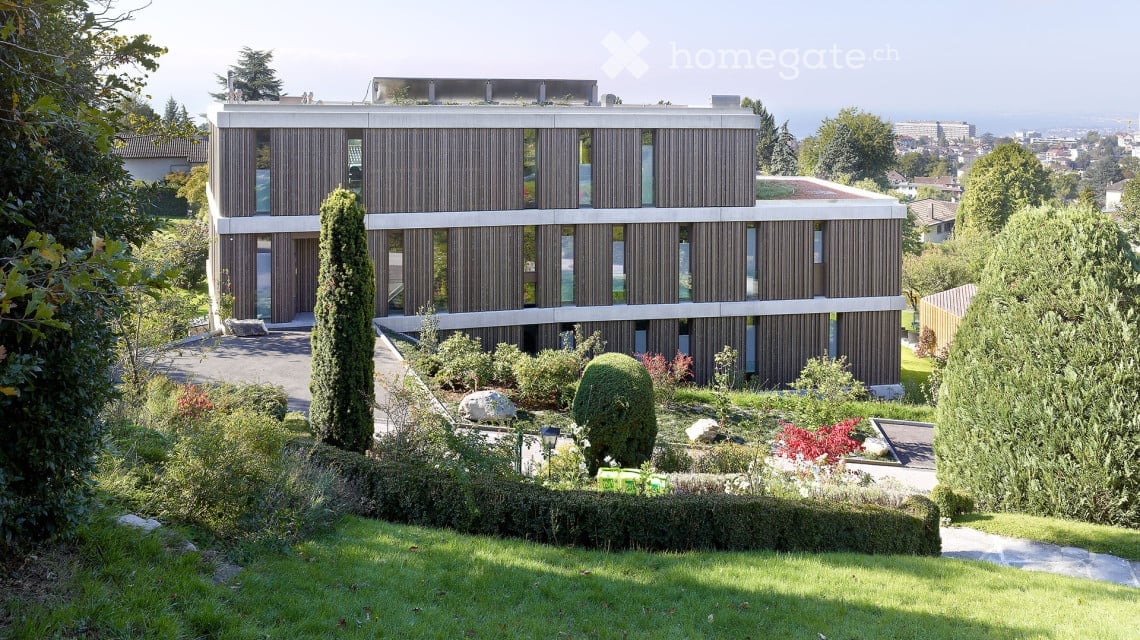 Modern wooden and white building, 4 stories, many windows, garden, green landscape, trees, and bushes