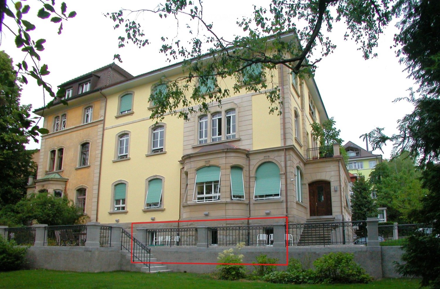 three story yellow house, green windows, two entrances, black metal fence, stairs leading up to main entrance, red line framing a portion of the house