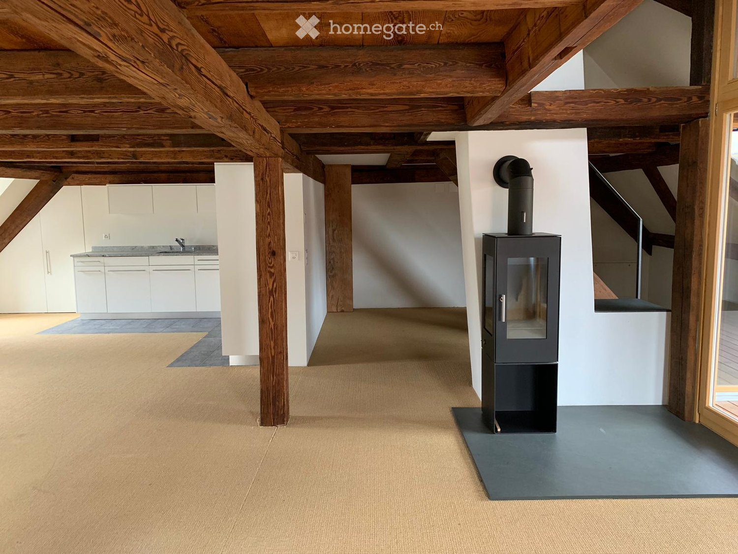 empty attic room with fireplace, wooden beams, glass window, kitchen and staircase in the background